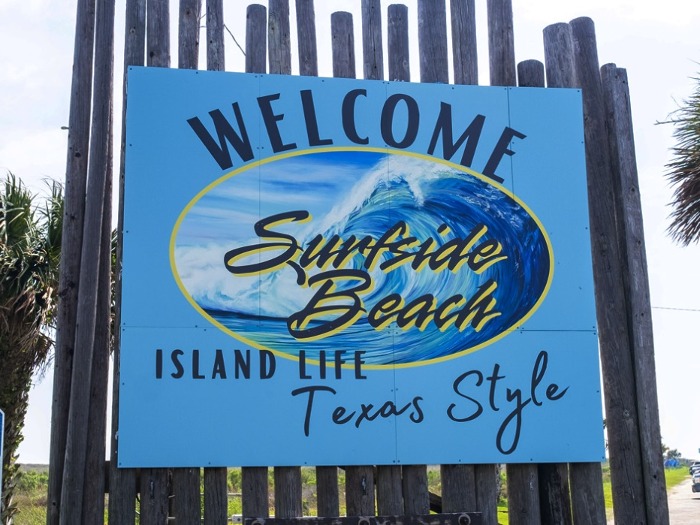 A blue outdoor sign reads, “Welcome to Surfside Beach. Island life, Texas style” mounted on wooden posts