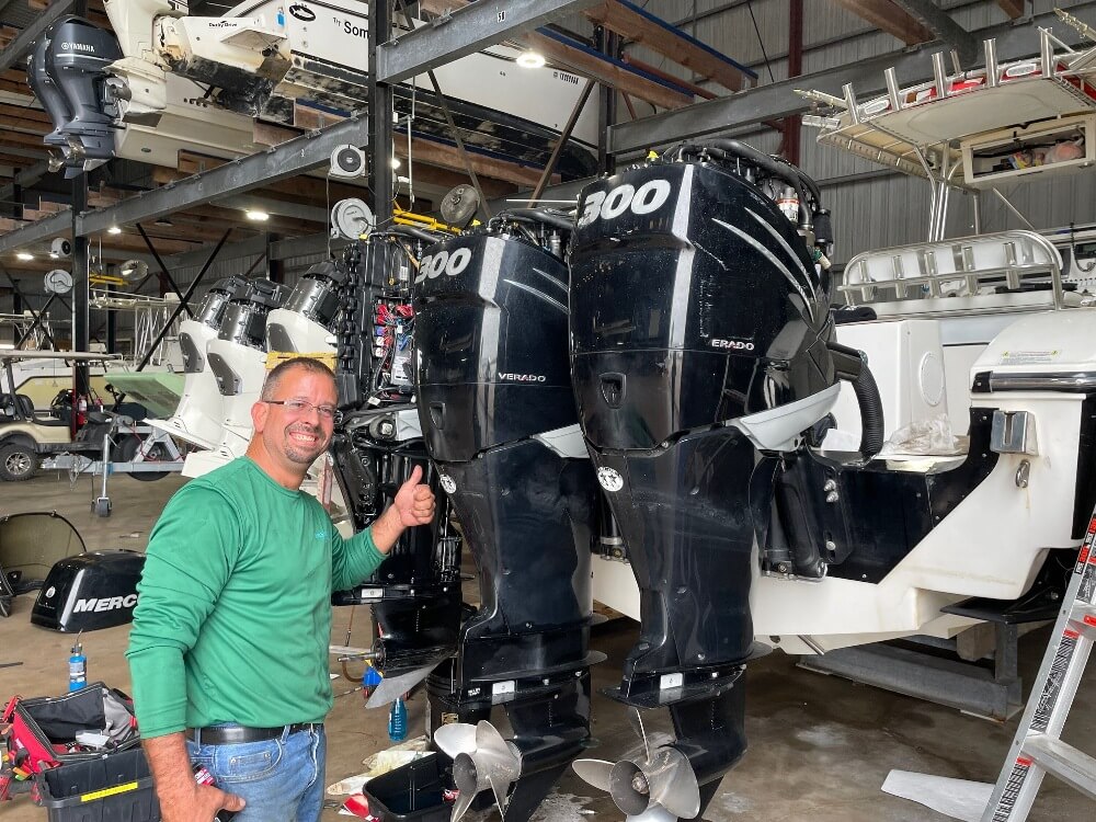 A man in a green shirt gives the camera a thumbs-up in front of two black outboard motor engines.